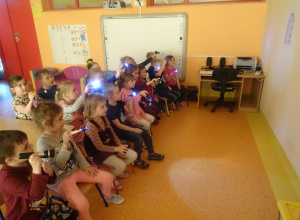 Grupa dzieci siedzi na krzesłach, trzymają włączone latarki skierowane na ścianę.